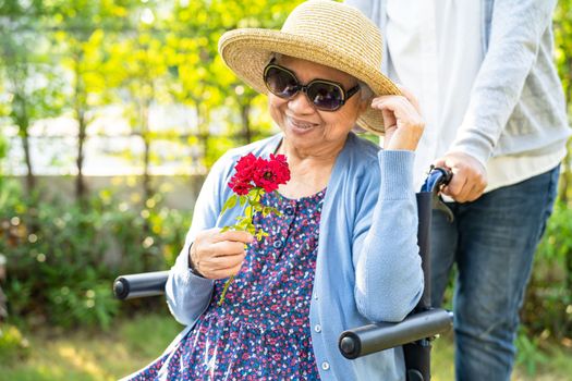 Caregiver daughter hug and help Asian senior or elderly old lady woman holding red rose on wheelchair in park.