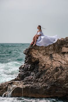 A woman in a storm sits on a stone in the sea. Dressed in a white long dress, waves crash against the rocks and white spray rises