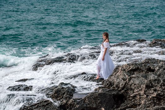 A woman stands on a rock in the sea during a storm. Dressed in a white long dress, the waves break on the rocks and white spray rises