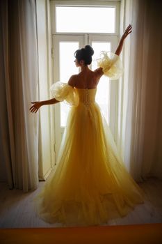 A woman's silhouette in a golden luxurious dress against the background of a window holds a curtain with her hands. Elegant lady in a yellow long silk dress with bare back, back view