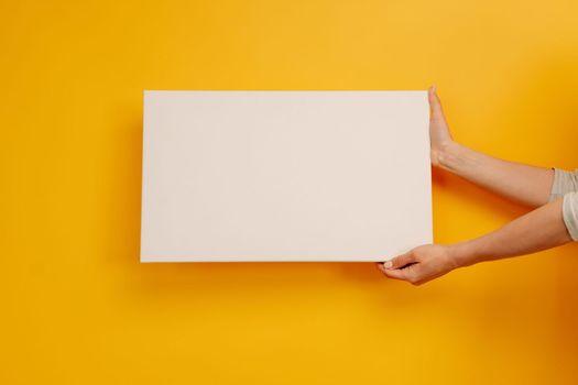 Woman hands holding white canvas paper blank with copyspace on yellow background. Empty sheet banner with place for text. Concept of creativity and new idea