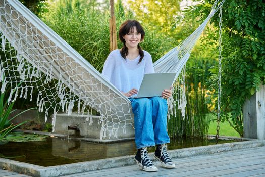 Teenage girl relaxing in hammock using laptop for leisure study, looking at camera. Adolescence, students, high school, technology, lifestyle, youth concept