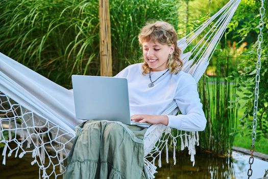 Teenage girl relaxing in hammock using laptop for leisure study. Adolescence, students, high school, technology, lifestyle, youth concept