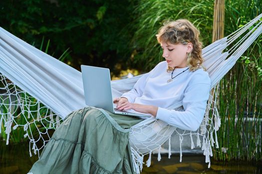 Teenage girl relaxing in hammock using laptop for leisure study. Adolescence, students, high school, technology, lifestyle, youth concept