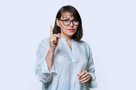 Middle aged serious woman showing index finger up, looking at camera, attention sign, on white studio background