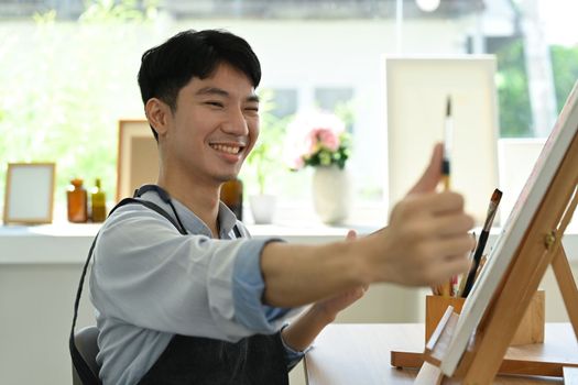 Happy asian male artist holding paintbrushes and palette painting on canvas at art studio. Education, hobby, art concept.