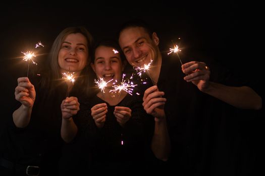 Happy family in black - woman, man and little girl, with a sparkler in hand the New Year's Christmas at home. The girl at the father on hands. New year 2023