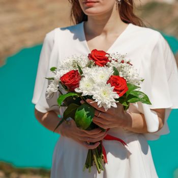 a bouquet of white chrysanthemums and red roses in the hands of the bride against the background of an azure lake