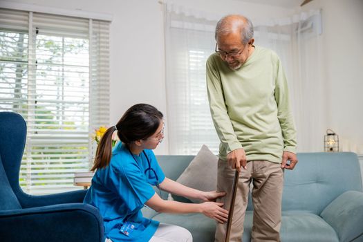 Asian young woman nurse checking knee and leg after surgery of senior old man patient suffering from pain in knee, doctor asking elderly man about pain symptom with walking stick