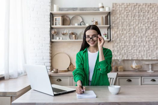 A young female student, a freelancer studies, works online, remotely at home from a laptop. She sits at the table, looks at the camera, holds his glasses, writes in a notebook, smiles.