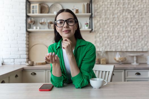 A young beautiful female blogger is sitting at home in the kitchen and talking online to the camera, explaining with her hands, smiling.