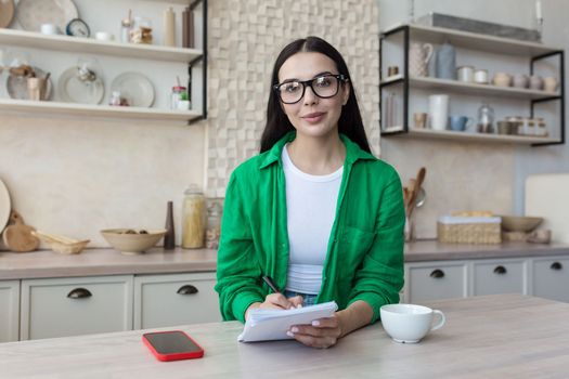 Portrait of a young woman teacher in glasses sitting at home in the kitchen in front of the camera. Works from home online. He is holding a notebook with a pen in his hands, smiling.