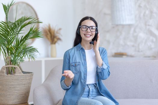 Distance Learning. Portrait of a young female teacher in glasses sitting at home on the sofa in a headset, talking and explaining to the camera. Works, teaches from home online.