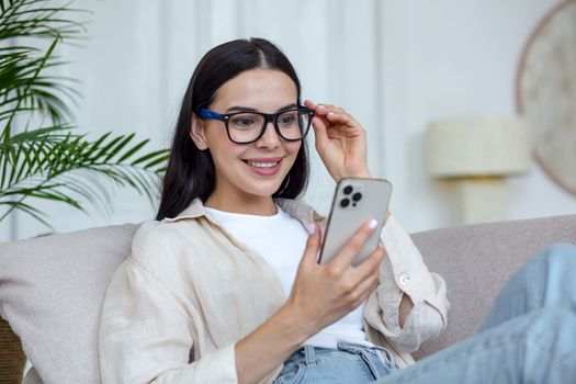 A happy young woman in quarantine lies on the couch at home and communicates with friends, parents, relatives through a video call from the phone. He smiles, says hello, holds his glasses in his hand.