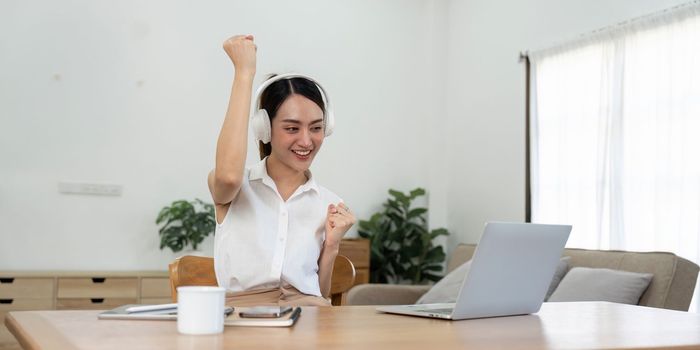 Asian teenage girl wearing headphones listening music while studying online on laptop, happy female student celebrating success sitting on floor in living room at home.