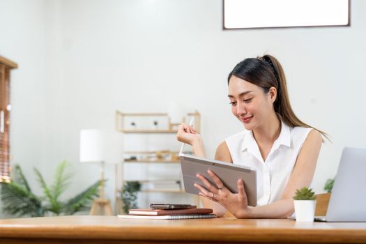 Portrait of young asian woman holding stylus pen while working on tablet computer at home.