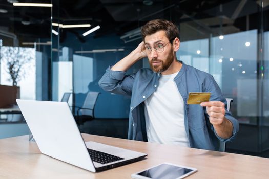 Bankruptcy, financial problems, unsuccessful online shopping. Shocked young man sitting in the office, holding a credit card, looking at a laptop. Checks the account, scratches the back of the head.