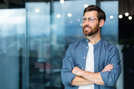 Portrait of a young successful man, businessman, freelancer, designer standing in the office, crossing his arms, confidently looking to the side