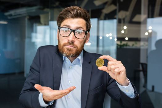 Close-up photo. Confused young business man holds a gold coin, cryptocurrency, bitcoin in his hand. He looks into the camera, points to a coin, points with his hand, asks a question.