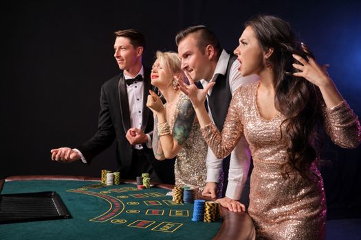 Side shot of a stylish rich friends playing poker at casino. Youth have lose. They are looking disappointed standing at the table against a red and blue backlights on black background. Risky gambling entertainment.