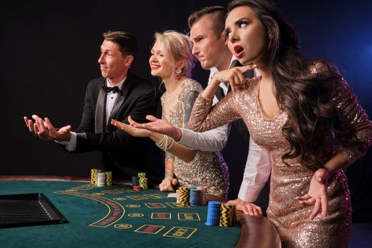 Side shot of a good-looking friends playing poker at casino. Youth have lose. They are looking upset standing at the table against a red and blue backlights on black background. Risky gambling entertainment.
