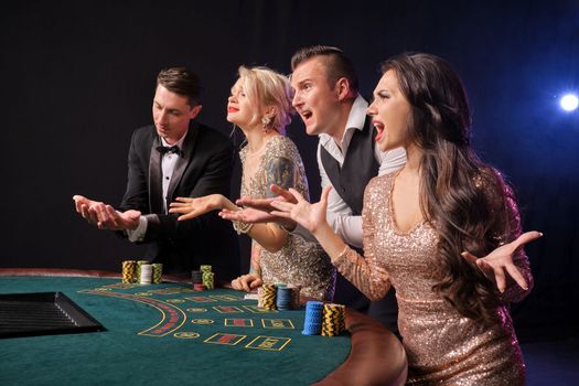 Side shot of a confused rich companions playing poker at casino. Youth have lose. They are screaming standing at the table against a red and blue backlights on black background. Risky gambling entertainment.