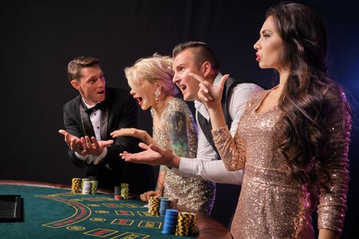 Side shot of a confused rich colleagues playing poker at casino. Youth have lose. They are looking unsatisfied standing at the table against a red and blue backlights on black background. Risky gambling entertainment.