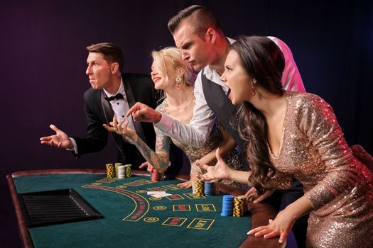 Side shot of a confused rich classmates playing poker at casino. Youth have lose. They are standing at the table against a red and blue backlights on black background. Risky gambling entertainment.