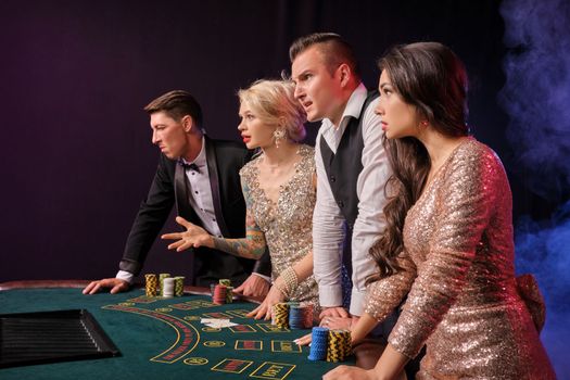 Side shot of a disappointed rich buddies playing poker at casino in smoke. Youth have lose. They are looking unhappy standing at the table against a red and blue backlights on black background. Risky gambling entertainment.