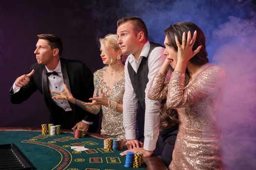 Side shot of a disappointed rich companions playing poker at casino in smoke. Youth have lose. They are looking confused standing at the table against a red and blue backlights on black background. Risky gambling entertainment.