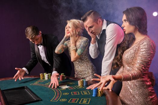Side shot of a disappointed rich colleagues playing poker at casino in smoke. Youth have lose. They are standing at the table against a red and blue backlights on black background. Risky gambling entertainment.