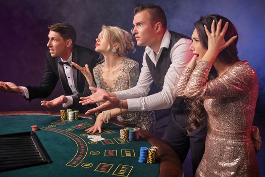 Side shot of a disappointed rich classmates playing poker at casino in smoke. Youth have lose. They are looking upset standing at the table against a red and blue backlights on black background. Risky gambling entertainment.