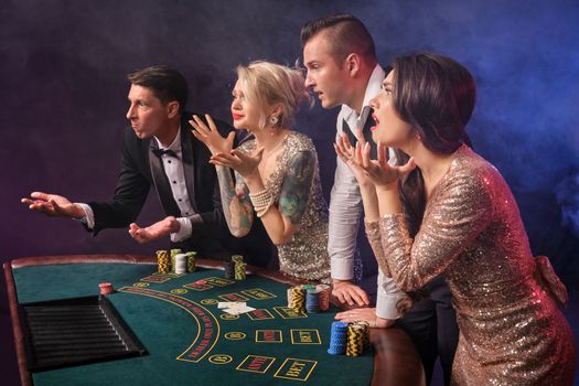 Side shot of a disappointed rich friends playing poker at casino in smoke. Youth have lose. They are standing at the table against a red and blue backlights on black background. Risky gambling entertainment.