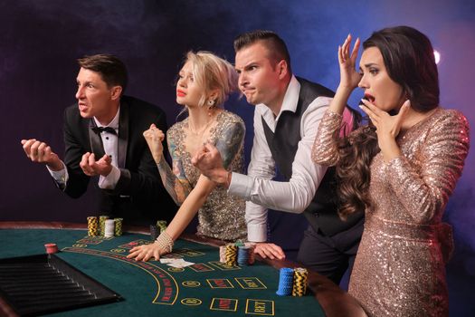 Side shot of an upset rich buddies playing poker at casino in smoke. Youth are making bets waiting for a big win. They are looking confused standing at the table against a red and blue backlights on black background. Risky gambling entertainment.