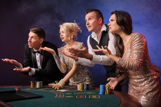 Side shot of an upset rich colleagues playing poker at casino in smoke. Youth are making bets waiting for a big win. They are looking unhappy standing at the table against a red and blue backlights on black background. Risky gambling entertainment.