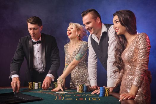 Side shot of a rejoicing rich classmates playing poker at casino in smoke. Youth are making bets waiting for a big win. They are standing at the table against a red and blue backlights on black background. Risky gambling entertainment.