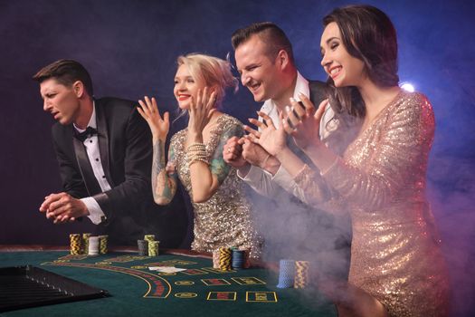 Side shot of an overjoyed rich buddies playing poker at casino in smoke. Youth are making bets waiting for a big win. They are clapping their hands standing at the table against a red and blue backlights on black background. Risky gambling entertainment.