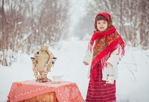 Adorable girl in old clothes in winter forest with a samovar and bagels