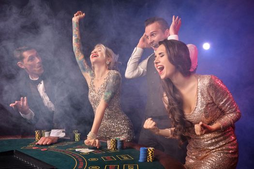 Side shot of an overjoyed rich colleagues playing poker at casino in smoke. Youth are making bets waiting for a big win. They are looking overjoyed standing at the table against a red and blue backlights on black background. Risky gambling entertainment.