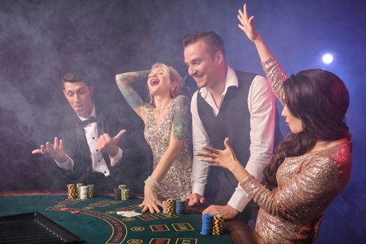 Side shot of an overjoyed rich friends playing poker at casino in smoke. Youth are making bets waiting for a big win. They are looking very happy standing at the table against a red and blue backlights on black background. Risky gambling entertainment.