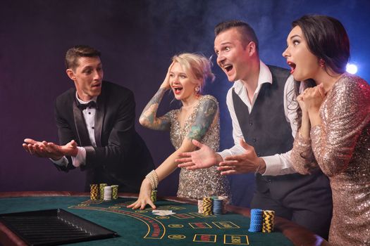 Side shot of a joyful rich friends playing poker at casino in smoke. Youth are making bets waiting for a big win. They are looking wondered standing at the table against a red and blue backlights on black background. Risky gambling entertainment.
