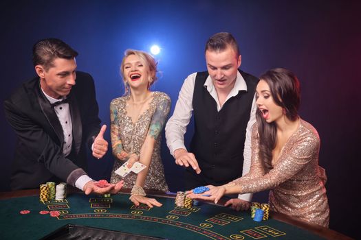 Group of a cheerful rich classmates are playing poker at casino. Youth are making bets waiting for a big win. They are having fun standing at the table against a red and blue backlights on black background. Risky gambling entertainment.