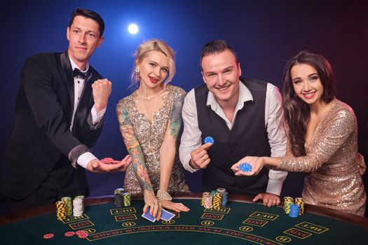 Group of an excited rich companions are playing poker at casino. Youth are making bets waiting for a big win. They are looking at the camera standing at the table against a red and blue backlights on black background. Risky gambling entertainment.