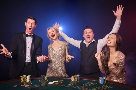 Group of an excited rich classmates are playing poker at casino. Youth are making bets waiting for a big win. They are standing at the table against a red and blue backlights on black background. Risky gambling entertainment.