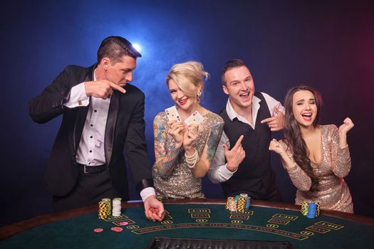 Group of an excited rich friends are playing poker at casino. Youth are making bets waiting for a big win. They are looking joyful standing at the table against a red and blue backlights on black background. Risky gambling entertainment.