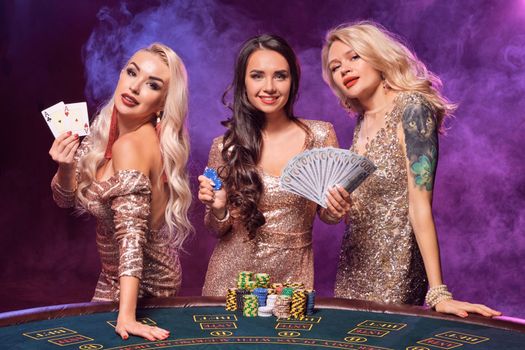 Charming females with a perfect hairstyles and bright make-up, dressed in a golden shiny dresses are posing standing at a gambling table. Poker concept on a black smoke background with pink and blue backlights. Casino.