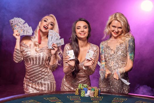 Charming women with a perfect hairstyles and bright make-up, dressed in a golden shiny dresses are posing standing at a gambling table and looking happy. Poker concept on a black smoke background with pink and blue backlights. Casino.