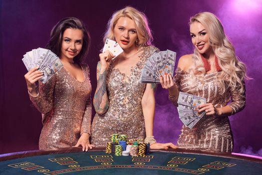 Beautiful ladies with a perfect hairstyles and bright make-up, dressed in a golden shiny dresses are posing standing at a gambling table and looking at the camera. Poker concept on a black smoke background with pink and blue backlights. Casino.