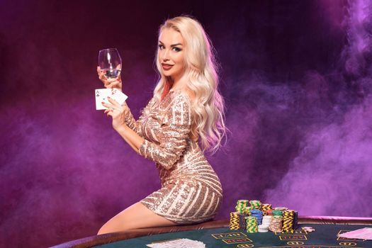 Close-up shot of a gorgeous girl with blond hair and bright make-up, dressed in a golden striped shiny dress, posing with playing cards and glass of champagne in her hands while sitting on a table with chips on it and smiling. Poker concept on a black smoke background with pink and blue backlights. Casino.