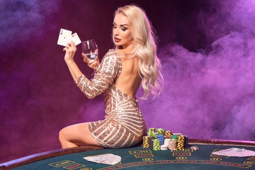 Close-up shot of a charming lady with blond hair and bright make-up, dressed in a golden striped shiny dress, posing with playing cards and glass of champagne in her hands while sitting on a table with chips on it. Poker concept on a black smoke background with pink and blue backlights. Casino.